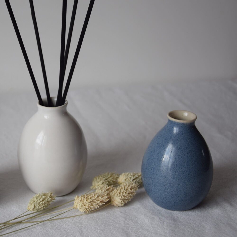 #CE123_MABL_3 Deux vases ambiance goutte d'eau en céramique côte à côte. L'un est blanc brillant agrémenté de cinq bâtonnets de rotin couleur noir, l'autre est bleu horizon. Entre les vases sont posés quelques épis de graminées séchés, montrant les utilisations pour ce petit vase en céramique soit en décoration soit comme contenant pour parfum d'ambiance en ajoutant les bâtonnets de rotin.