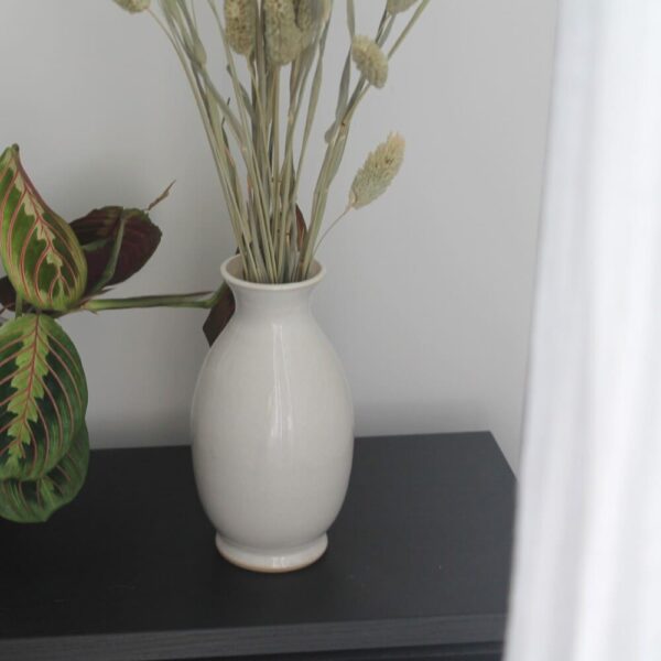 #CE100_BLA Le vase Onésime contient un bouquet d'épis séchés et est posé sur un meuble noir contre un mur blanc. Coté droit on voit une partie d'un voilage blanc et coté gauche apparaissent quelques feuilles d'une plante verte dont les feuilles sont dans un dégradé de verts et les veinures de la feuilles se dessinent en rouge.