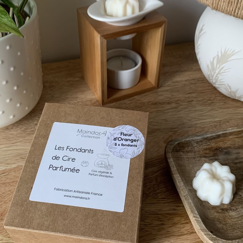 MaindorA-Coffret 8 fondants est fermé. On voit bien l'étiquette commerciale avec le nom "les fondants de cire parfumée" avec un dessin représentant un fondant et un brûle-parfum en céramique avec le message "cire végétale et parfum d'exception ! Une étiquette ronde est apposée dessus en quinconce dans un coin avec inscrit le nom du parfum "Fleur d'oranger, 8 fondants". La boite kraft est posée sur un meuble en bois dans une ambiance très apaisante.
