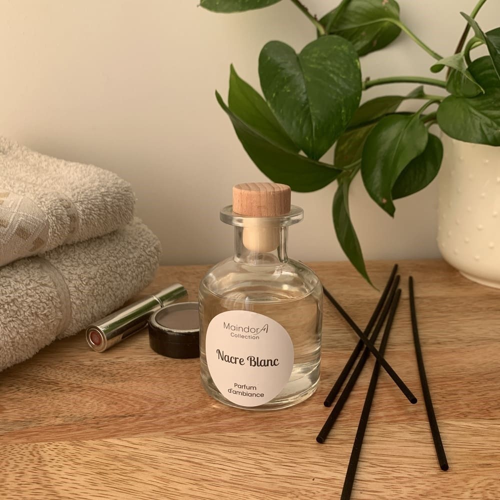 MaindorA-Diffuseur APOTHICAIRE transparent dans une salle de bain à coté d'une pile de serviettes éponges écrues pliées, des accessoires de maquillage disposés ci et là, une plante verte et des bâtonnets de diffusion noirs posé sur la commode en bois.