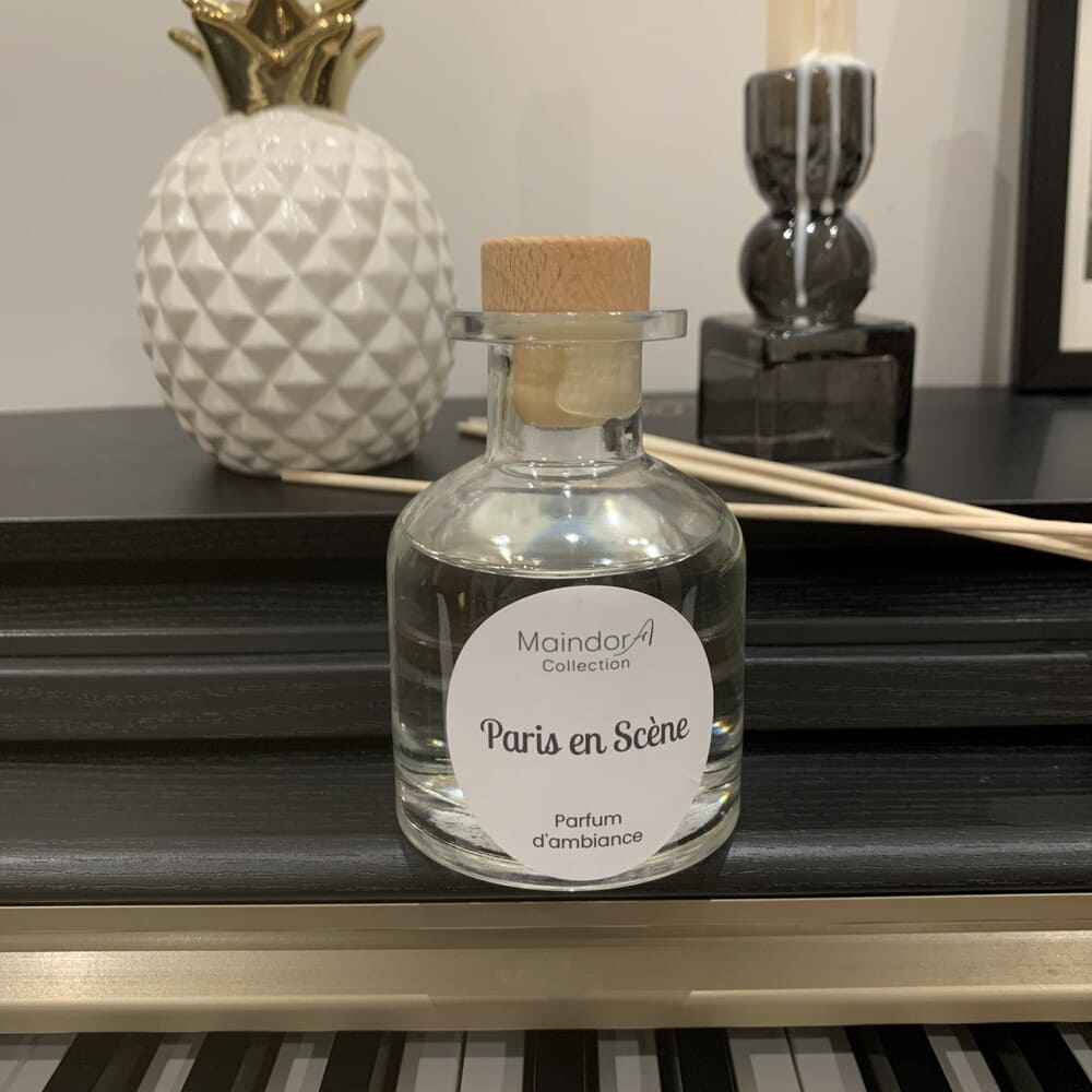 MaindorA-le Diffuseur de parfum d'ambiance PARIS EN SCENE, présenté dans son flacon APOTHICAIRE transparent a fière allure posé sur le bord d'un piano Noir. des tiges en rotin naturel viennent rappeler le bouchon en bois clair du diffuseur.