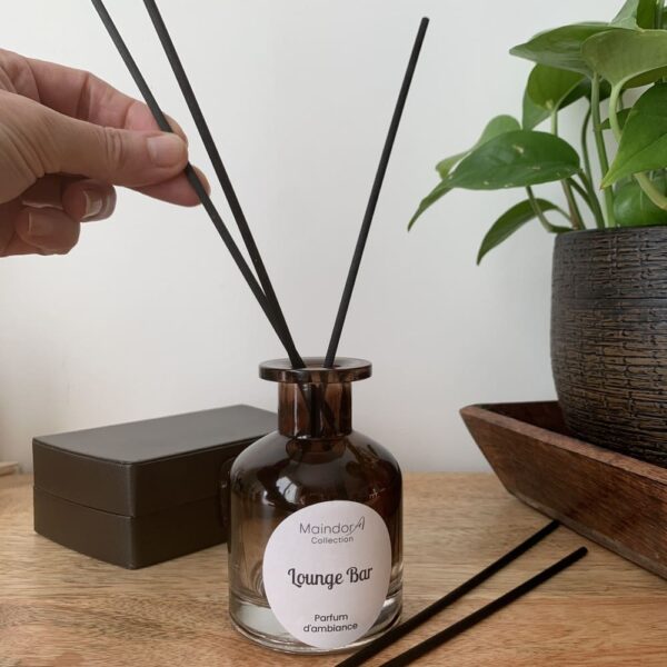 MaindorA-une main insère délicatement les bâtonnets de diffusion noirs à l'intérieur du Diffuseur de parfum d'ambiance, parfum LOUNGE BAR. Le flacon apothicaire couleur ambre fumé, est posé sur son meuble en bois à coté une boîte en cuir brun et de l'autre un plateau en bois foncé accueille un pot en terre marron et noire avec une de plante verte. Une décoration très chaleureuse et mystérieuse.