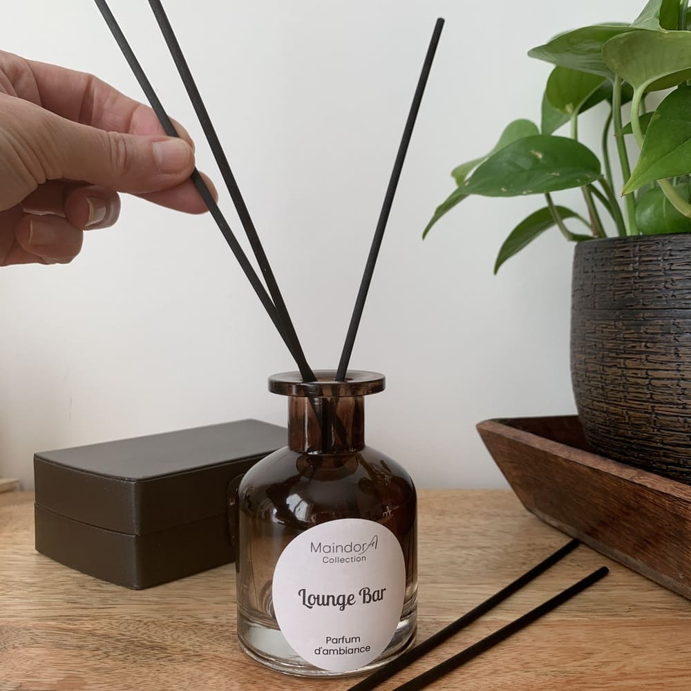MaindorA-une main insère délicatement les bâtonnets de diffusion noirs à l'intérieur du Diffuseur de parfum d'ambiance, parfum LOUNGE BAR. Le flacon apothicaire couleur ambre fumé, est posé sur son meuble en bois à coté une boîte en cuir brun et de l'autre un plateau en bois foncé accueille un pot en terre marron et noire avec une de plante verte. Une décoration très chaleureuse et mystérieuse.