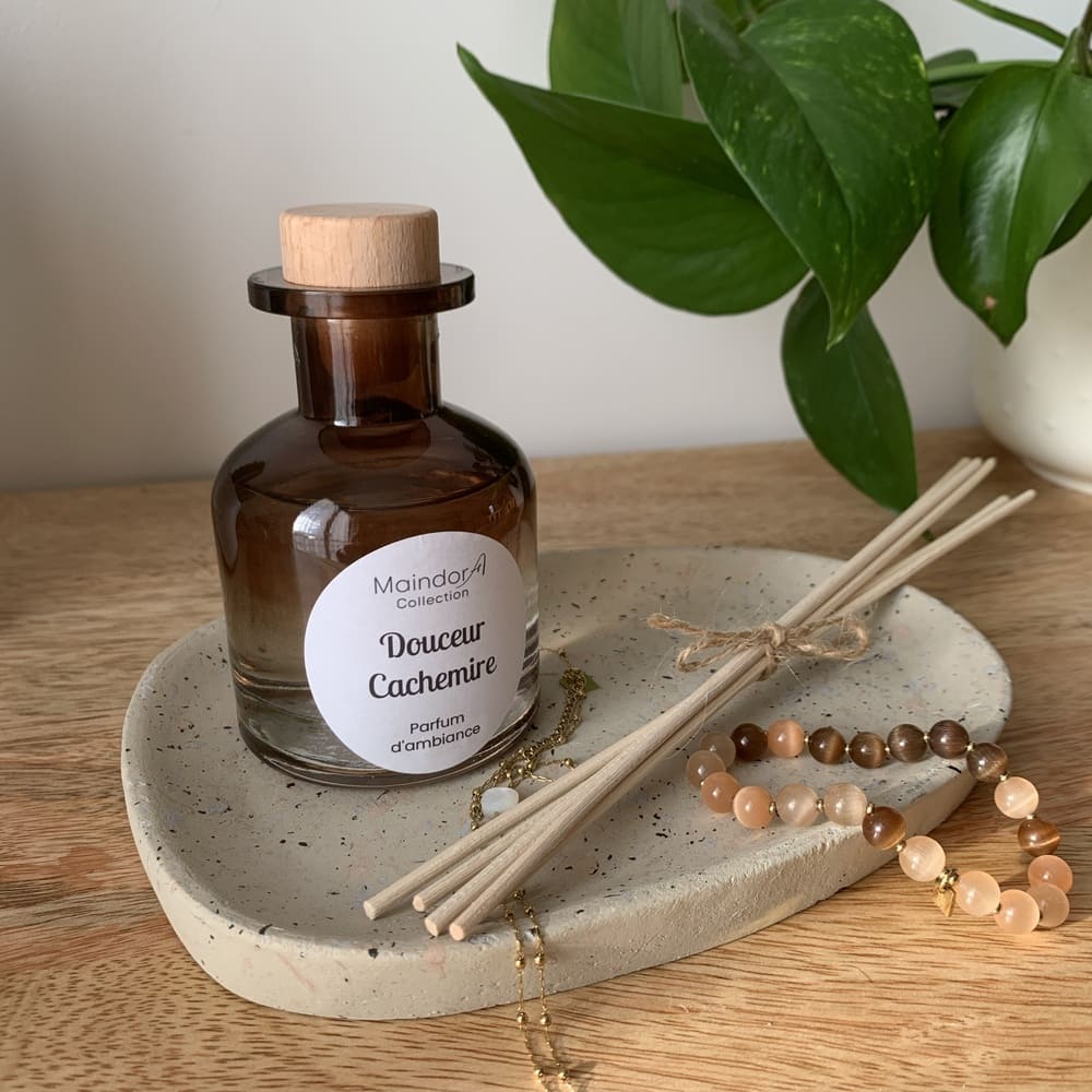 MaindorA-Le diffuseur modèle APOTHICAIRE ambre fumé avec son bouchon en bois clair est délicatement posé dans un plateau ovale en pierre écru et moucheté de points noirs. Des bijoux trainent sur celui-ci ainsi que le bouquet de tiges de rotin nature reliées par un brin en lin. Un ensemble plein de douceur comme son parfum d'ambiance DOUCEUR CACHEMIRE;