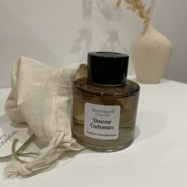 MaindorA- Diffuseur de parfum d'ambiance DOUCEUR CACHEMIRE. Un petit flacon tout arrondit couleur ambrée et bouchon noir. Il est sur l'image comme à peine sorti de son pochon en lin. Dans le fond de l'image un peu flouté un petit vase en grès brut écru contient des brins naturels séchés.