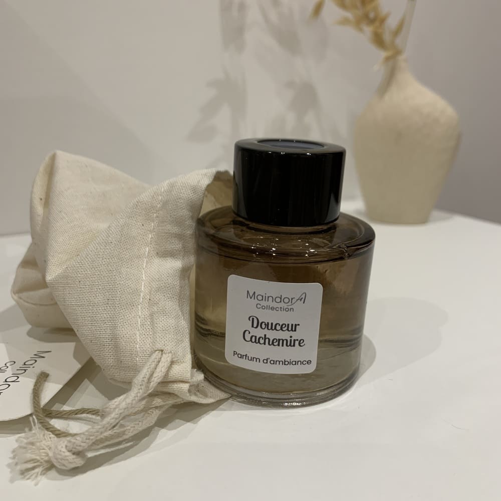 MaindorA- Diffuseur de parfum d'ambiance DOUCEUR CACHEMIRE. Un petit flacon tout arrondit couleur ambrée et bouchon noir. Il est sur l'image comme à peine sorti de son pochon en lin. Dans le fond de l'image un peu flouté un petit vase en grès brut écru contient des brins naturels séchés.