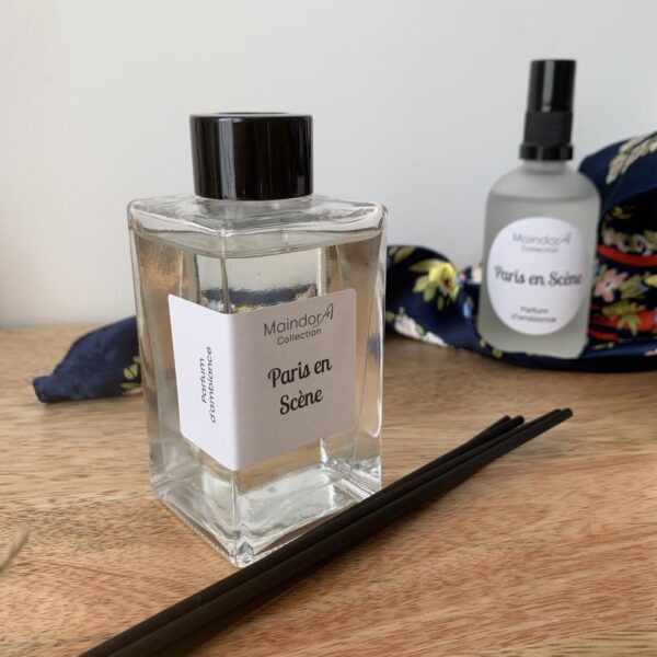 MaindorA-Diffuseur parfum d'ambiance modèle Singulier cristal clair contenant le parfum PARIS EN SCENE. Autour de lui ses bâtonnets noirs posés à coté et un peu dans le fond un spray de parfum d'ambiance contenant la même senteur est installé sur un foulard en soie sur fond bleu marine