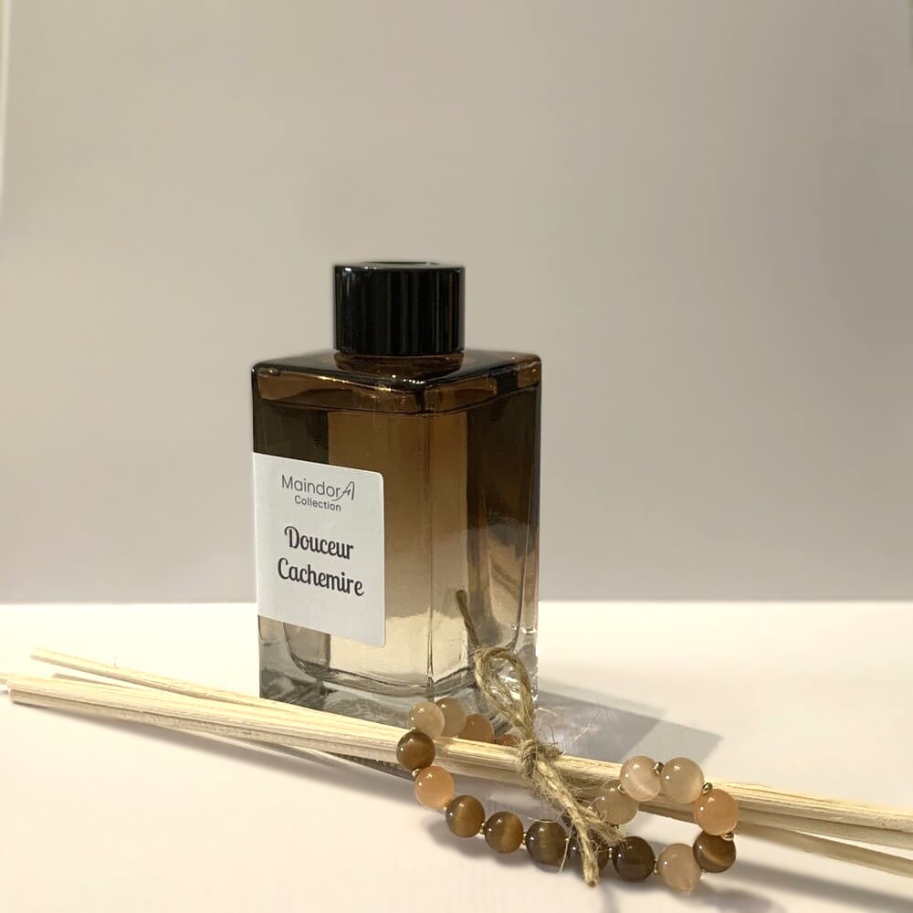 MaindorA-Posé sur une table blanche, s présente le Diffuseur de parfum d'Ambiance modèle SINGULIER. Sa couleur Ambre fumé apporte beaucoup de douceur dans les reflets du flacon rectiligne. à coté se trouve un bouquet de tiges en rotin naturel maintenu par une cordelette en chanvre. un bracelet de perles ambres, nacres et quartz roses traîne à coté du diffuseur rappelant les mêmes teintes ambrées.