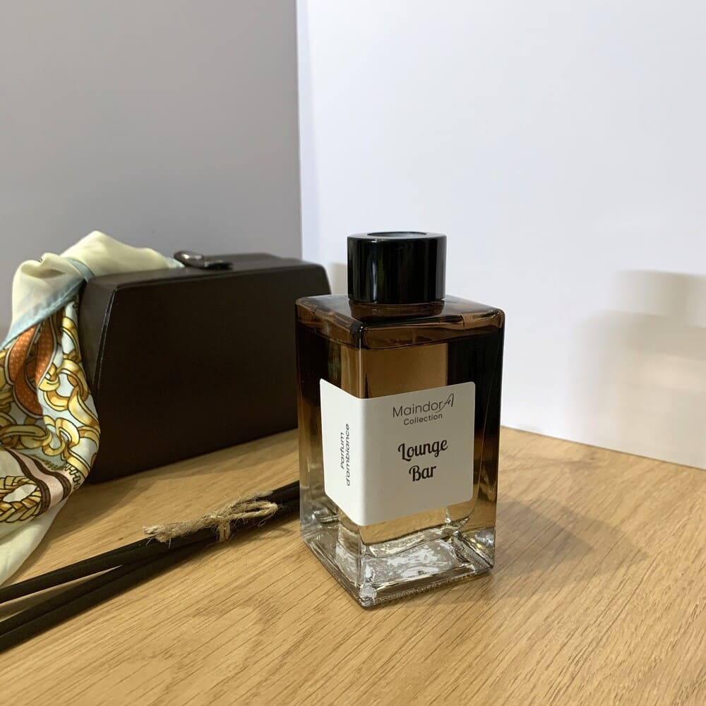 MaindorA-Diffuseur de parfum d'Ambiance couleur Ambre dégradé dans un flacon rectangulaire. Bouchon noir et bâtonnets noirs. Posé sur une table en bois, un étuis en cuir brun laissant dépasser un foulard de soie très chic. Le parfum inscrit sur l'étiquette est "LOUNGE BAR" pour une Ambiance tamisée et enveloppante.
