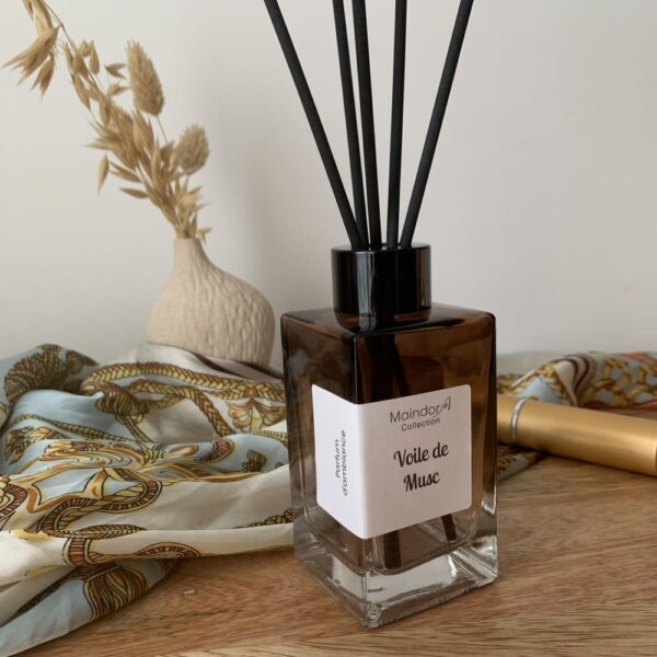 MaindorA-Diffuseur modèle SINGULIER couleur Ambre dégradé avec capuchon noir et tiges en fibre noires. Forme rectangulaire posé sur un meuble en bois à coté un tube de rouge à lèvres, un foulard en soie imprimée avec des motifs ocres et bleu ciel et blanc, un petit vase en grés brut motifs Mishima et des brins séchés dedans. Le parfum d'ambiance est VOILE DE MUSC.
