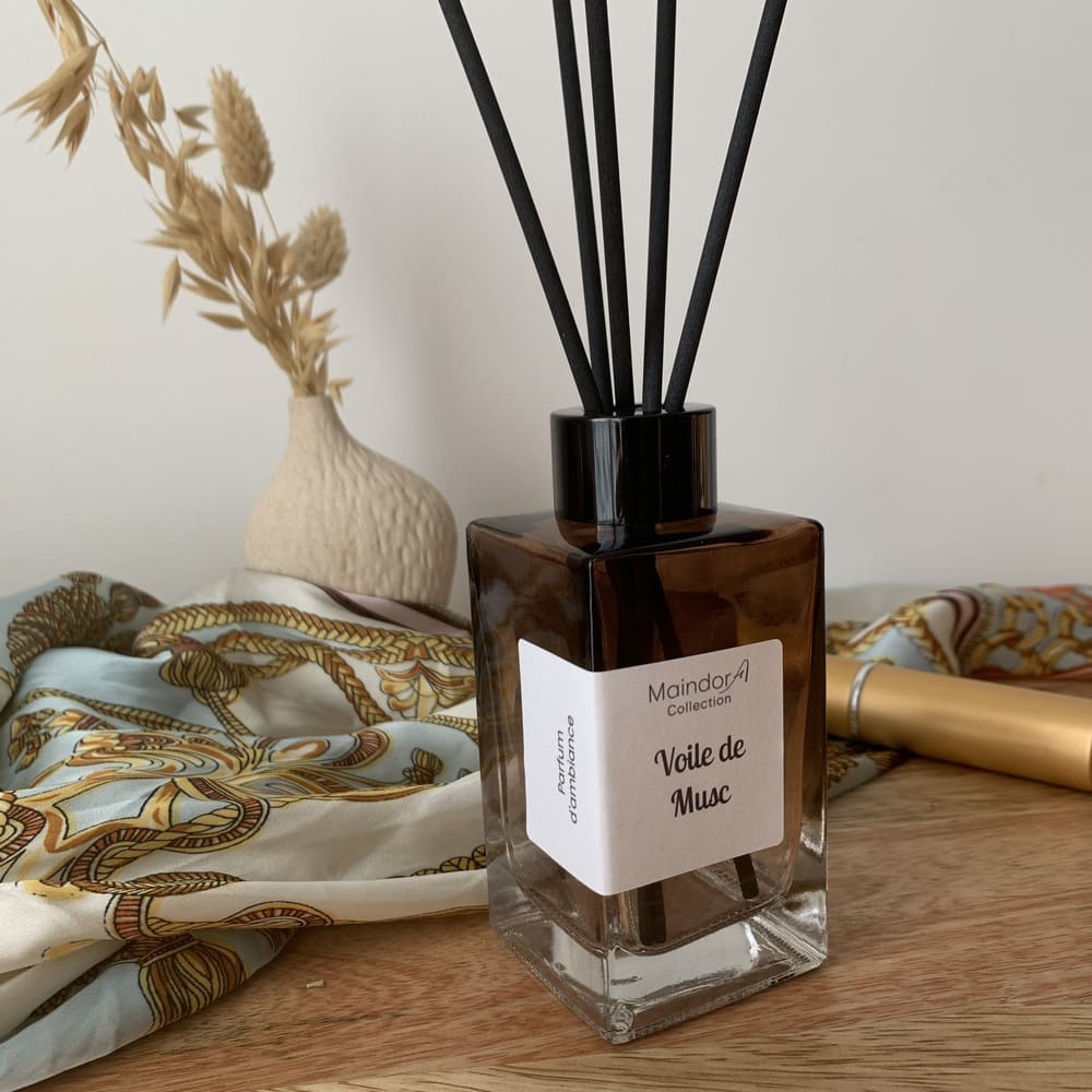 MaindorA-Diffuseur modèle SINGULIER couleur Ambre dégradé avec capuchon noir et tiges en fibre noires. Forme rectangulaire posé sur un meuble en bois à coté un tube de rouge à lèvres, un foulard en soie imprimée avec des motifs ocres et bleu ciel et blanc, un petit vase en grés brut motifs Mishima et des brins séchés dedans. Le parfum d'ambiance est VOILE DE MUSC.