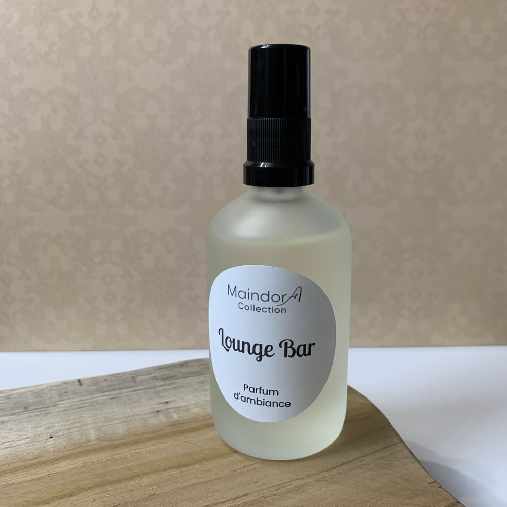 MaindorA-Flacon vaporisateur présenté ici sur un plateau en bois brut en plein écran. Le spray parfum d'intérieur dans un coloris un peu nacré, sa grande étiquette ronde indiquant le parfum d'ambiance "Lounge Bar" est mis en valeur dans cette déco.