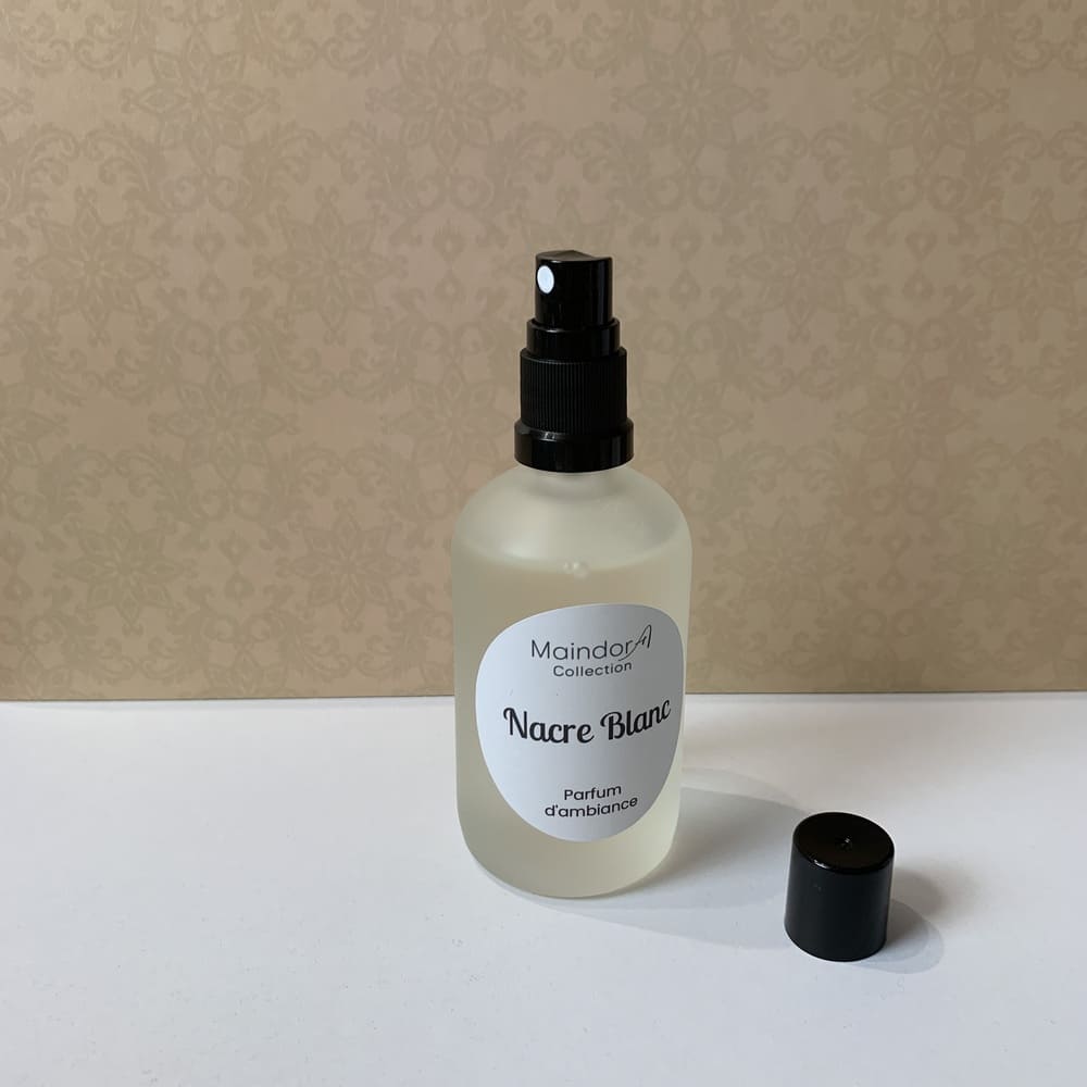 MaindorA-Flacon Spray de 100 ml posé sur un meuble blanc devant une tapisserie murale beige. il est en verre dépoli et son bouchon vaporisateur est noir ainsi que son capuchon d'ailleurs retiré et posé au pied du flacon. L'étiquette ronde au milieu de la bouteille indique la senteur NACRE BLANC - parfum d'ambiance.