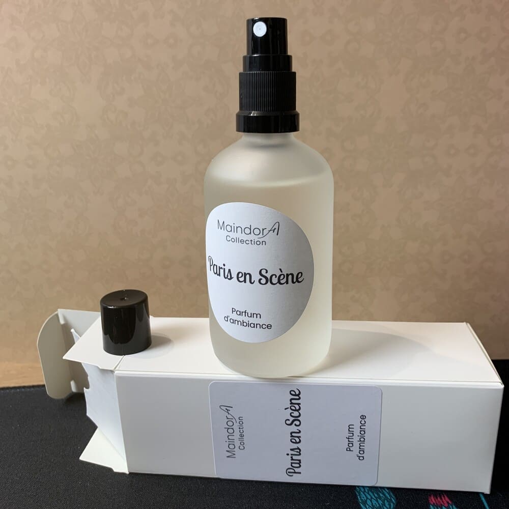 MaindorA-Flacon Spray parfum d'ambiance avec son capuchon noir ôté, est posé sur son étui cartonné blanc. A l'intérieur de cette boîte on distingue un insert cartonné démontrant une qualité pour ce produit conçu artisanalement dans l'atelier MaindorA. Le parfum d'ambiance représenté ici est Paris en scène que l'on retrouve également en version diffuseurs sur des modèle APOTHICAIRE, SINGULIER et DECOUVERTE le tout en coloris cristal clair.