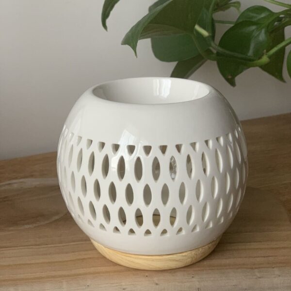 MaindorA-Coffret Fondants Parfumés ÉCLAT, présentation du brûle-parfum ici en grand plan. On voit bien la forme arrondie et d'un émail d'un blanc éclatant de la partie ajourée par des formes ovales alignées sur 3 rangées. En office de finition une ligne ajourée par de petites formes triangulaires au-dessus et en dessous des lignes des formes ovales. La forme de la coupelle est inséré au brûle-parfum dans un seul tenant de cette forme arrondi reposant sur un socle en céramique de couleur beige/jaune (sable).