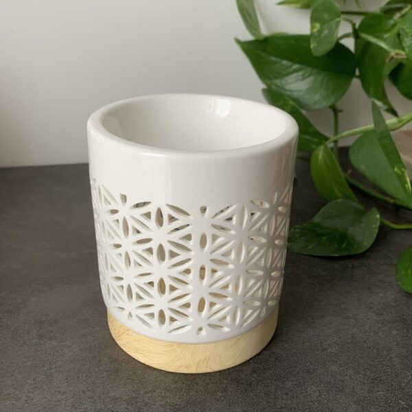 MaindorA-Coffret Fondants Parfumés FLEUR DE VIE avec le brûle parfum en céramique ici posé en gros plan sur un meuble gris. En arrière plan le feuillage d'une plante verte cela fait bien ressortir l'émail blanc de la partie ajourée du brule-parfum sur son socle beige/jaune.