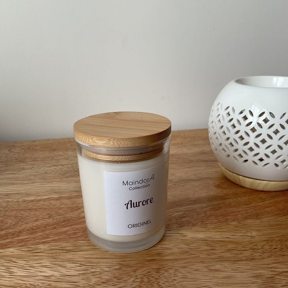MaindorA- Bougie Artisanale à la cire végétale, parfum Aurore posée sur un meuble en bois rappelant son couvercle en bambou. A coté un brûle-parfum rond blanc ajouré de motifs géométriques. l'ambiance est apaisante pleine authenticité .