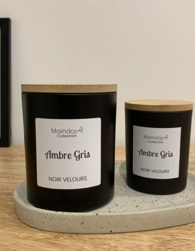 MaindorA - l'image montre les deux formats de bougies parfumées coulées à la main de la collection NOIR VELOURS. Les deux pots noirs avec leur couvercle en bambou sont posés sur un plateau en céramique dans une ambiance très sobre. On distingue parfaitement le parfum Ambre Gris indiqué sur les bougies.