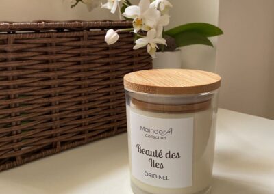 MaindorA-Bougie parfumée Artisanale Collection ORGINEL ici proposée avec le parfum BEAUTE DES ILES aux notes Notes olfactives : Florales, orangée et vanillée. La bougie porte son couvercle en bambou posée sur une table blanche, une grande boite tressée en osier en toile de fond à coté d'une belle orchidée aux fleurs blanches qui surplombent la bougie. Cette mise en scène inspire beaucoup de douceur l'esprit d'ailleurs de la collection de bougies ORIGINEL