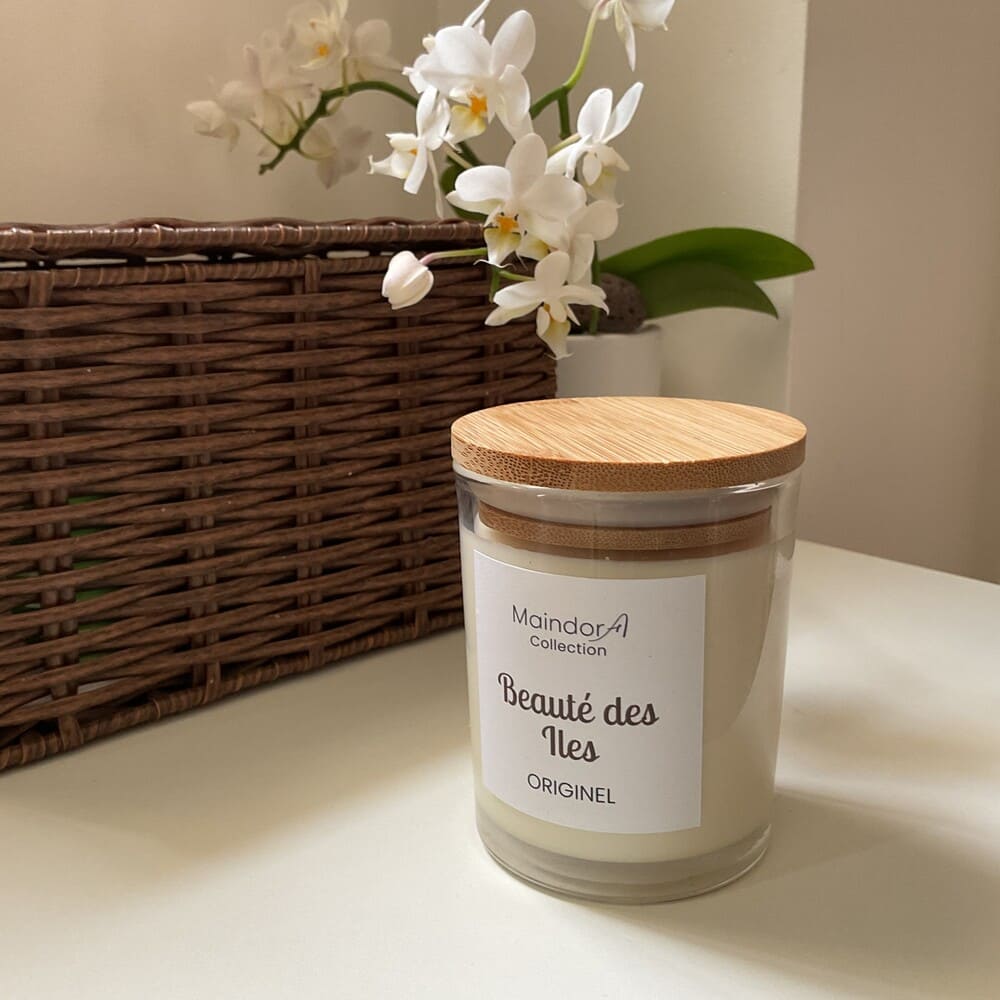 MaindorA-Bougie parfumée Artisanale Collection ORGINEL ici proposée avec le parfum BEAUTE DES ILES aux notes Notes olfactives : Florales, orangée et vanillée. La bougie porte son couvercle en bambou posée sur une table blanche, une grande boite tressée en osier en toile de fond à coté d'une belle orchidée aux fleurs blanches qui surplombent la bougie. Cette mise en scène inspire beaucoup de douceur l'esprit d'ailleurs de la collection de bougies ORIGINEL