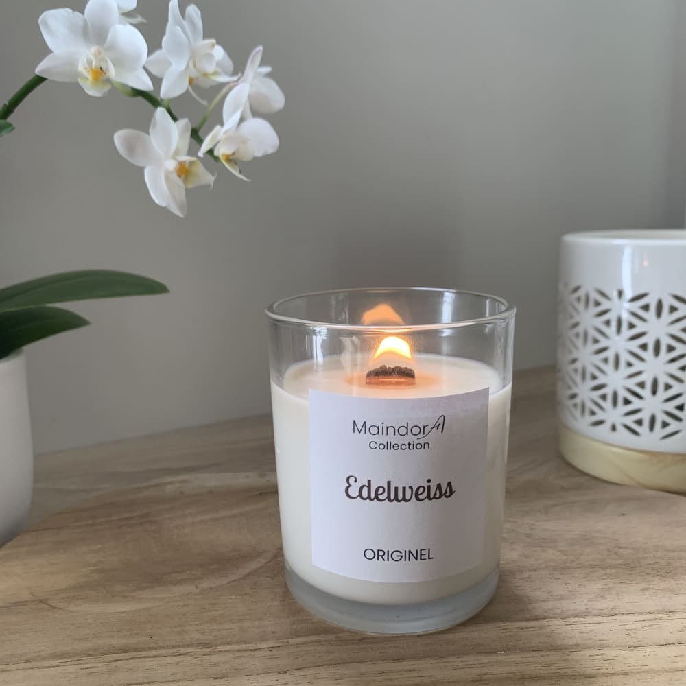 MaindorA- L'image présente la bougie parfumée artisanale dans un verre transparent qui permet de voir la mèche en bois de cerisier allumée. Tout autour les éléments créent une ambiance douce et chaleureuse comme le bois clair du plateur, un pot blanc avec orchidée à fleurs blanches à gauche, dans le coin droit un brule-parfum blanc ajouré aux motifs géométriques.