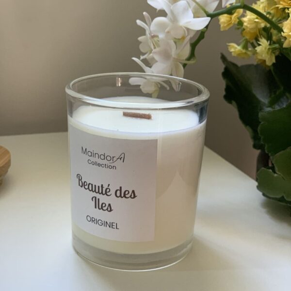 MaindorA-Bougie parfumée artisanale coulée à la main. Dans cette image elle est présentée sans son couvercle en bambou. On distingue bien la cire végétale et la mèche en bois de cerisier en son centre. en décor en arrière plan de coté quelques fleurs blanches et jaunes qui accentuent la douceur de la bougie.