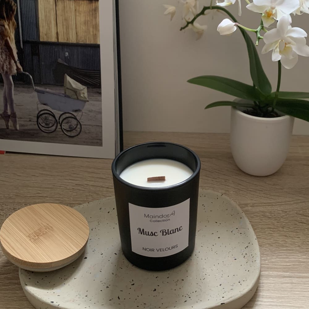 MaindorA-Bougie artisanale Parfumée Musc Blanc, une fragrance qui évoque la pureté et la sensualité. Dans cette image le pot noire de la bougie est posé sur un plateau en pierre écrue un peu moucheté. En arrière plan une orchidée blanche trône dans sont pot blanc. Sur la droite on voit un bout d'un grand livre photos aux couleurs très douces.