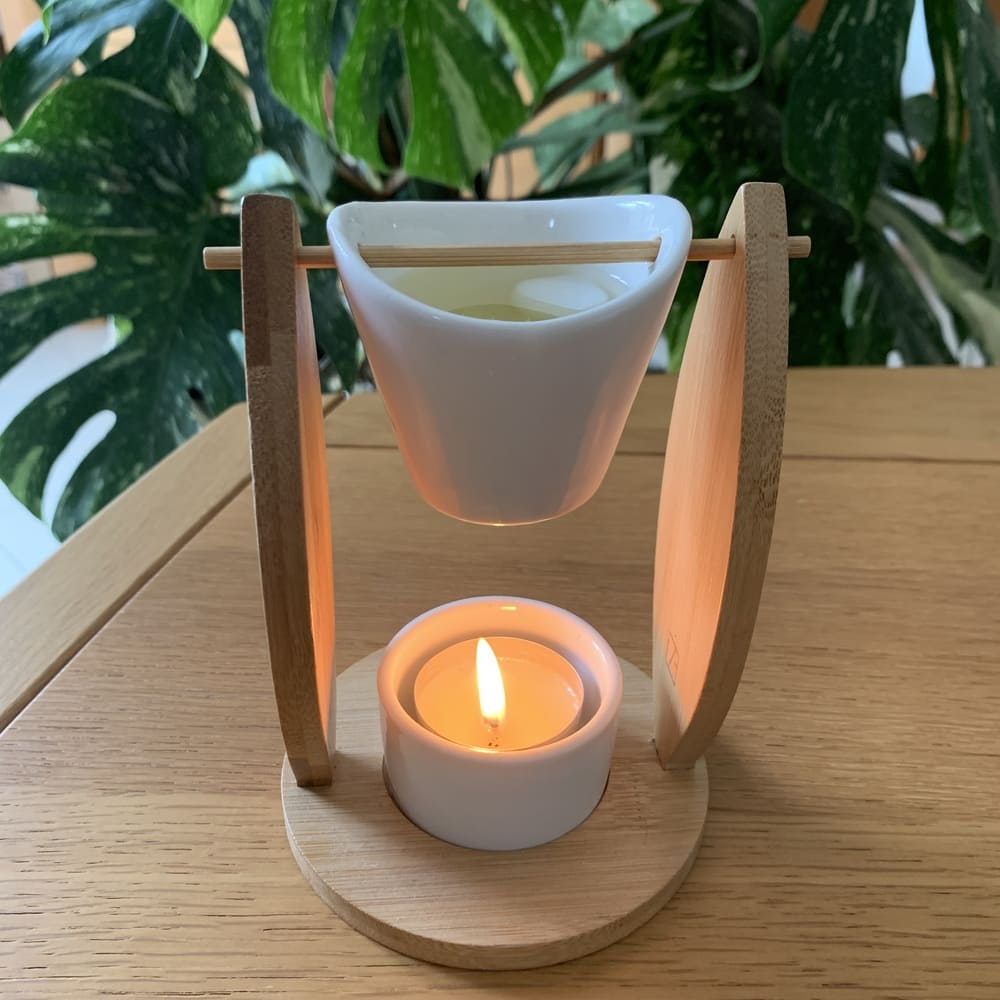 MaindorA-Ici le Brûle-parfum ALIZÉ mis en situation avec sa bougie chauffe-plat allumée qui a déjà bien fait fondre le fondant parfumé dans son seau en céramique. Il est posé sur une table en bois, Une grande plante monstera en arrière plan donne une atmosphère de douceur et de naturel.