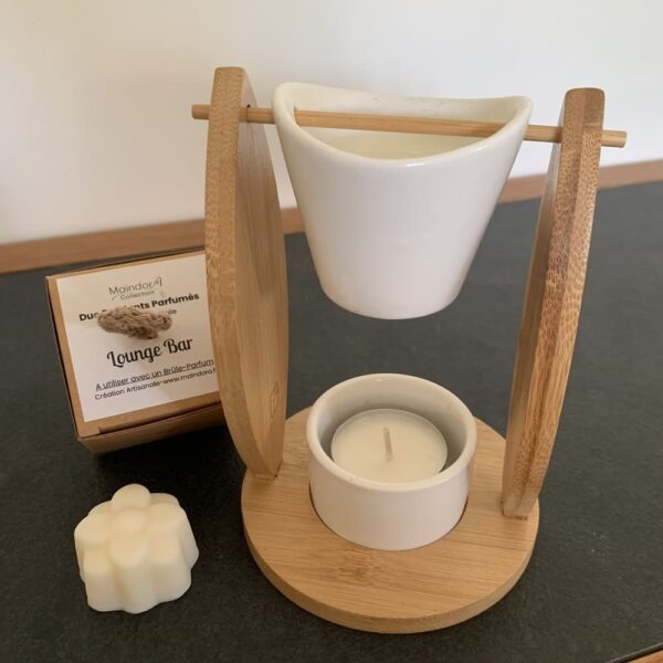 MaindorA-Dans cette image nous avons un gros plan sur le Brûle-parfum ALIZÉ en bambou et céramique blanche, accompagné d'un fondant parfumé et de sa boîte. Le seau en céramique blanche accueillant le galet de cire est suspendu par une tige en bambou. Cette tige passe de part en part à travers un trou situé aux extrémités hautes des deux cotés en forme de voile dont la base est fixée à un socle rond.