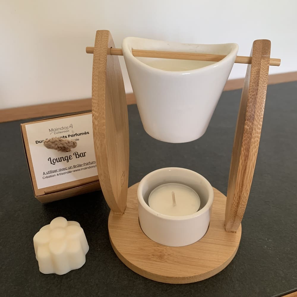 MaindorA-Dans cette image nous avons un gros plan sur le Brûle-parfum ALIZÉ en bambou et céramique blanche, accompagné d'un fondant parfumé et de sa boîte. Le seau en céramique blanche accueillant le galet de cire est suspendu par une tige en bambou. Cette tige passe de part en part à travers un trou situé aux extrémités hautes des deux cotés en forme de voile dont la base est fixée à un socle rond.