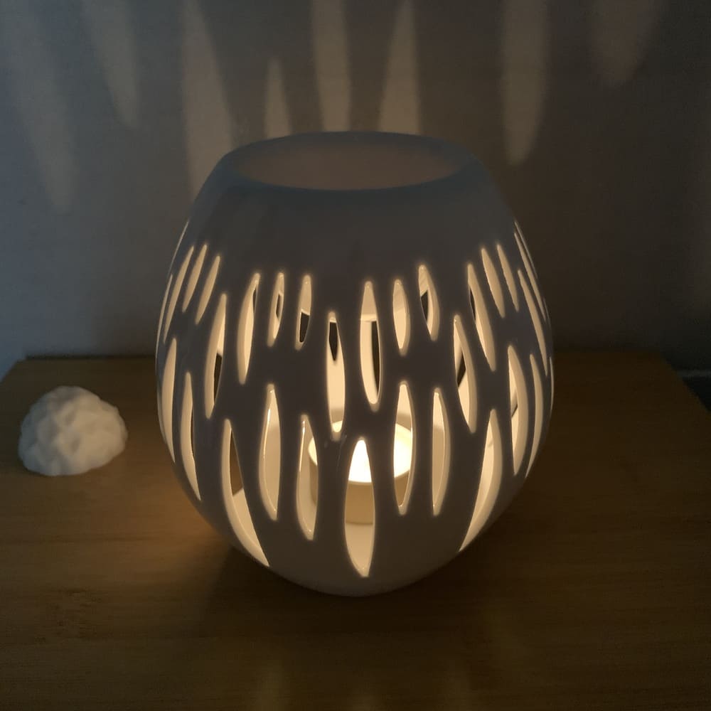 MaindorA-présentation du brûle-parfum NOVA allumé grâce à la bougie chauffe-plat. Pendant que le fondant parfumé fond délicatement dans la coupelle au dessus, la céramique ajourée du bruleur diffuse sa lumière à travers les motifs. Une ambiance particulière se créé bercée par les halos de lumière et la senteur qui se déploie dans la pièce.