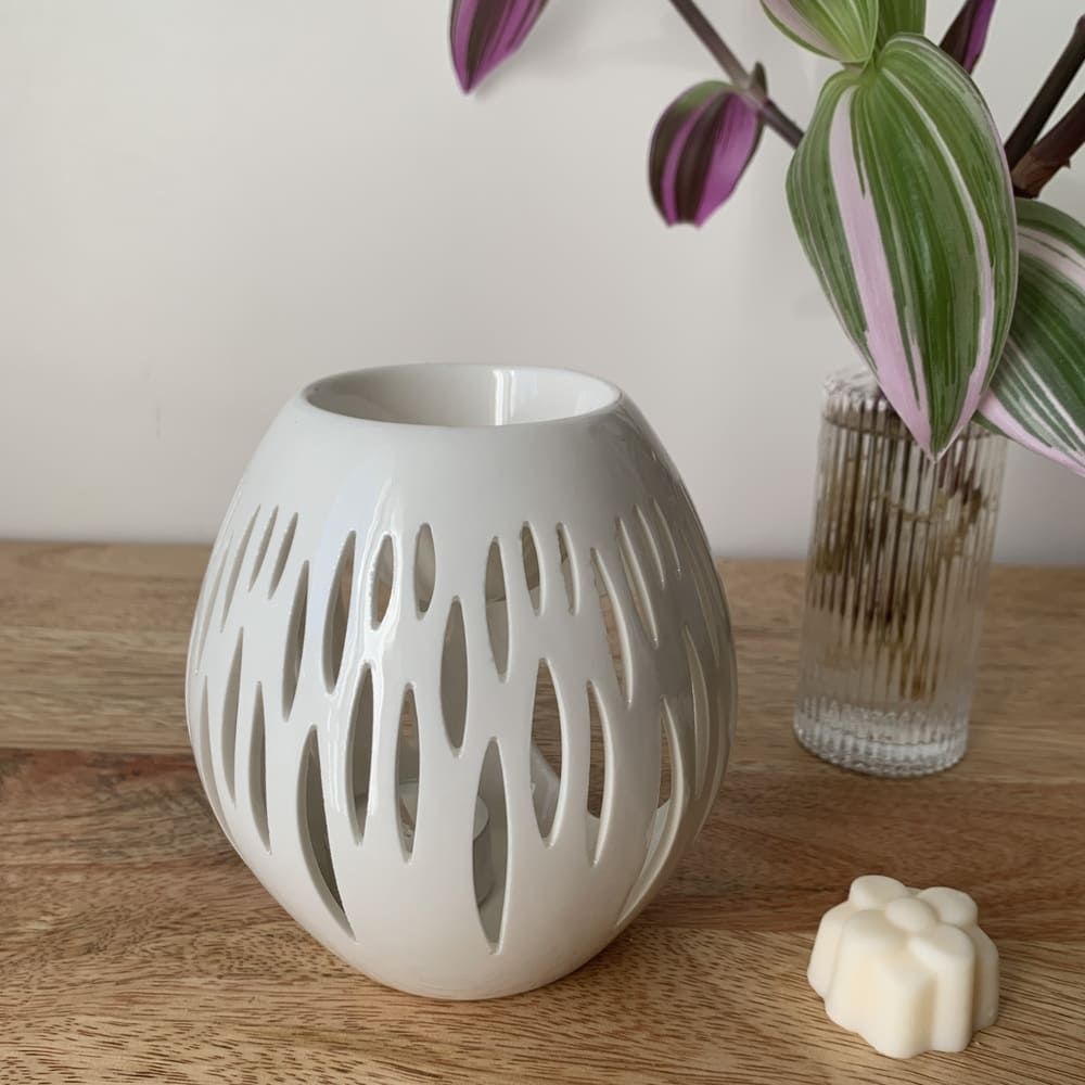 MaindorA-présentation du brûle-parfum NOVA tout en élégance grâce à sa céramique laquée blanche ajourée. Sa forme Ovale lui donne un air élancé. Son design s'intègre très bien dans tout intérieur. Ici le brûle -parfum NOVA est posé sur un meuble en bois naturel de manguier.